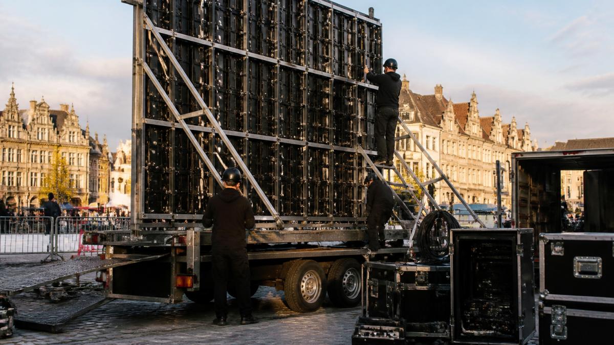 Technici bouwen een groot LED-scherm op een aanhangwagen op tijdens een buitenevenement in een Belgische stad, met bekabeling en een regieruimte zichtbaar in het late namiddaglicht